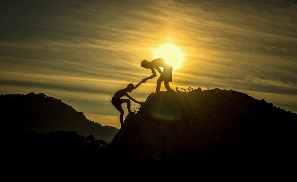one man reaching down to help another man up to to the top of a mountain at sunset